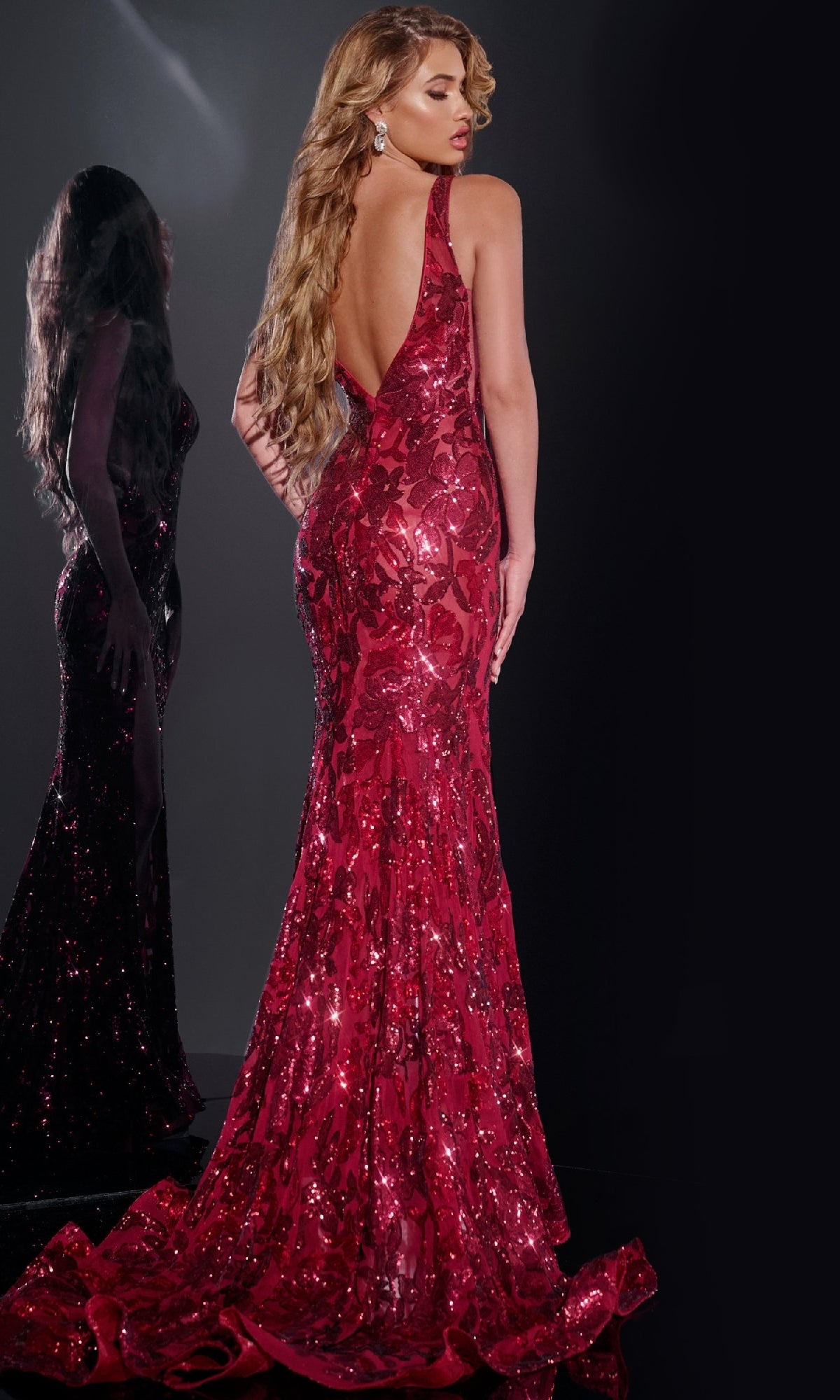 A woman with long wavy hair wears the Jovani 40755 Sleeveless Long Sequin Prom Dress in deep red, featuring a floor-length design and low back, as she stands before a mirror against a dark background.